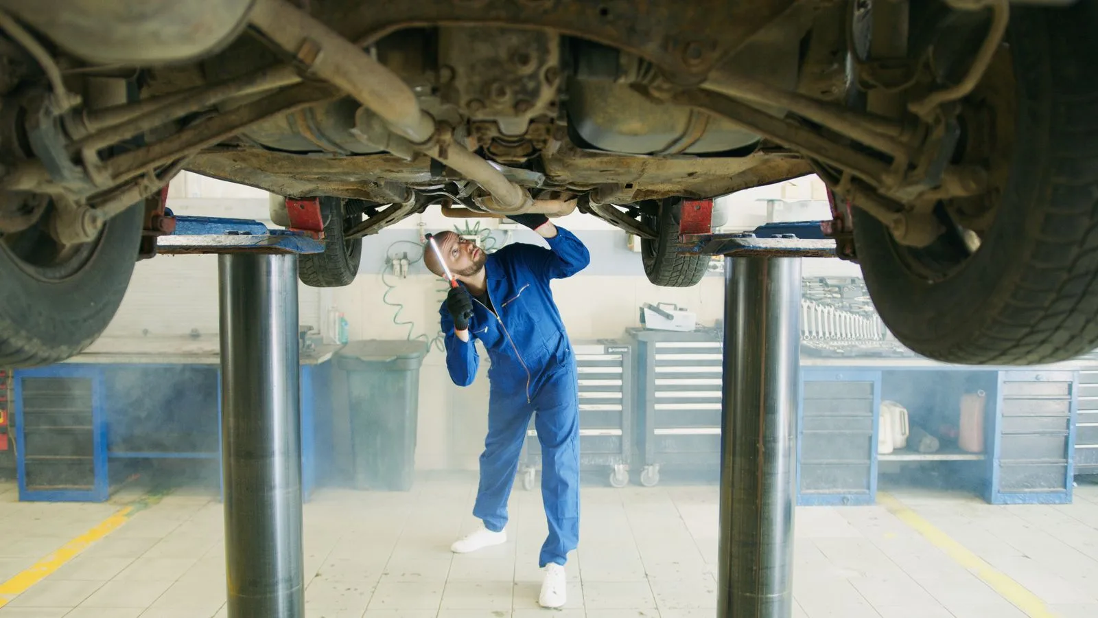 Mechanic in blue coveralls inspecting the underside of a car raised on a garage lift