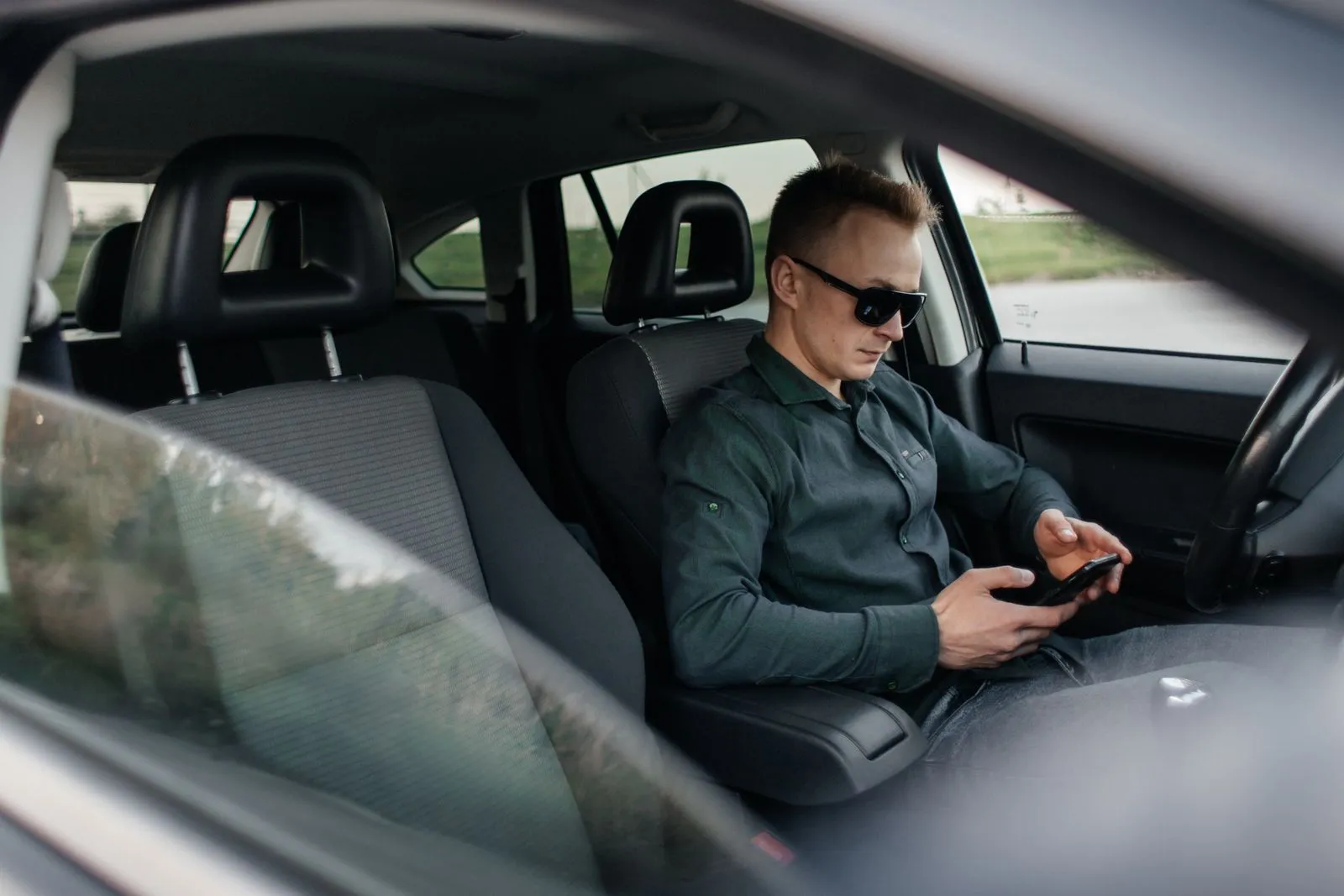 Man in sunglasses sitting in a car driver seat checking his phone, seen through the rear window