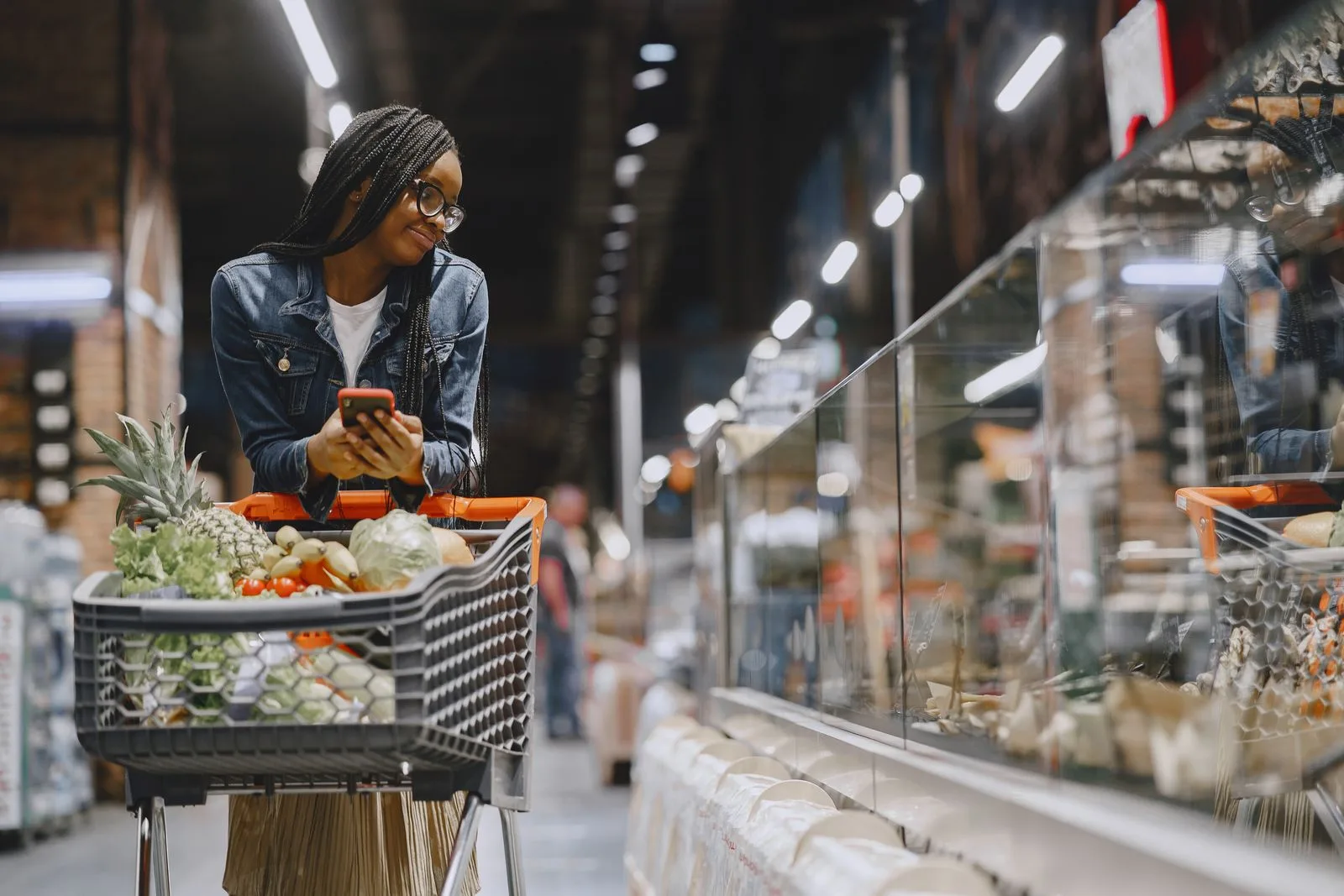 Shopper checking her phone while pushing a full grocery cart through a supermarket