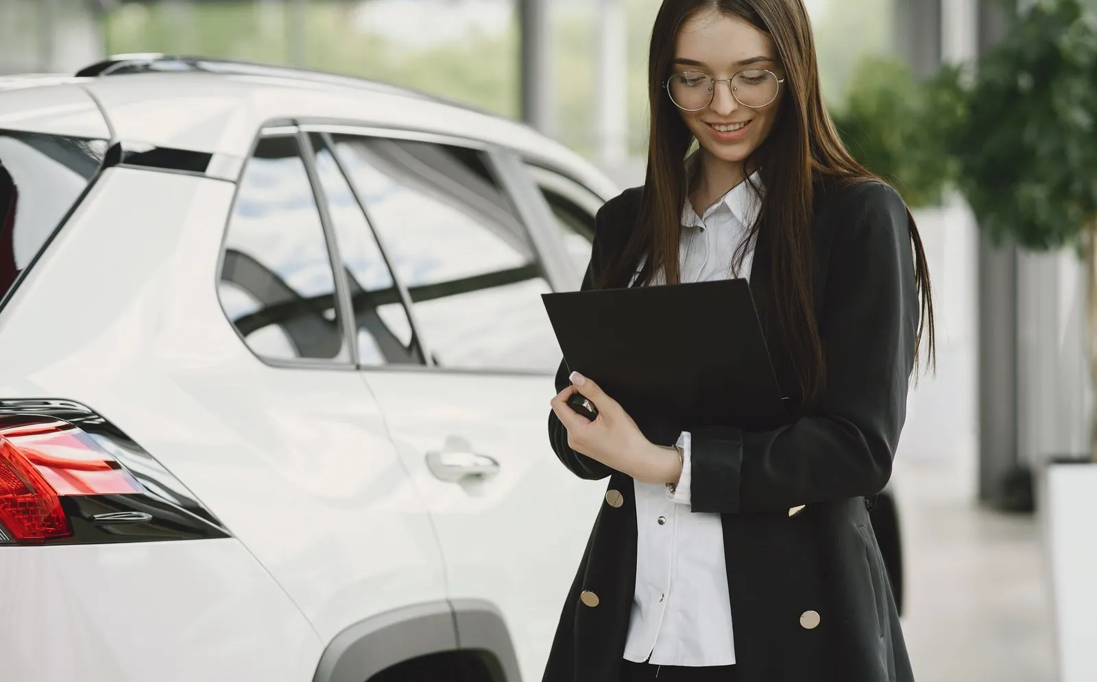 A woman reviewing paperwork next to a white SUV inside a car dealership showroom