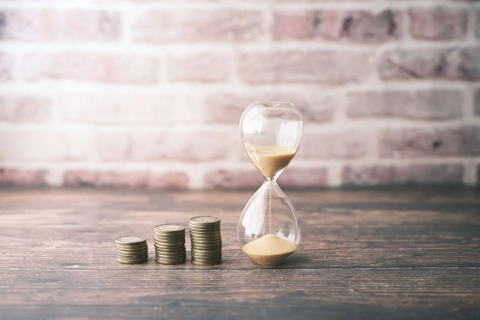 An hourglass beside three rising stacks of coins on a wooden table