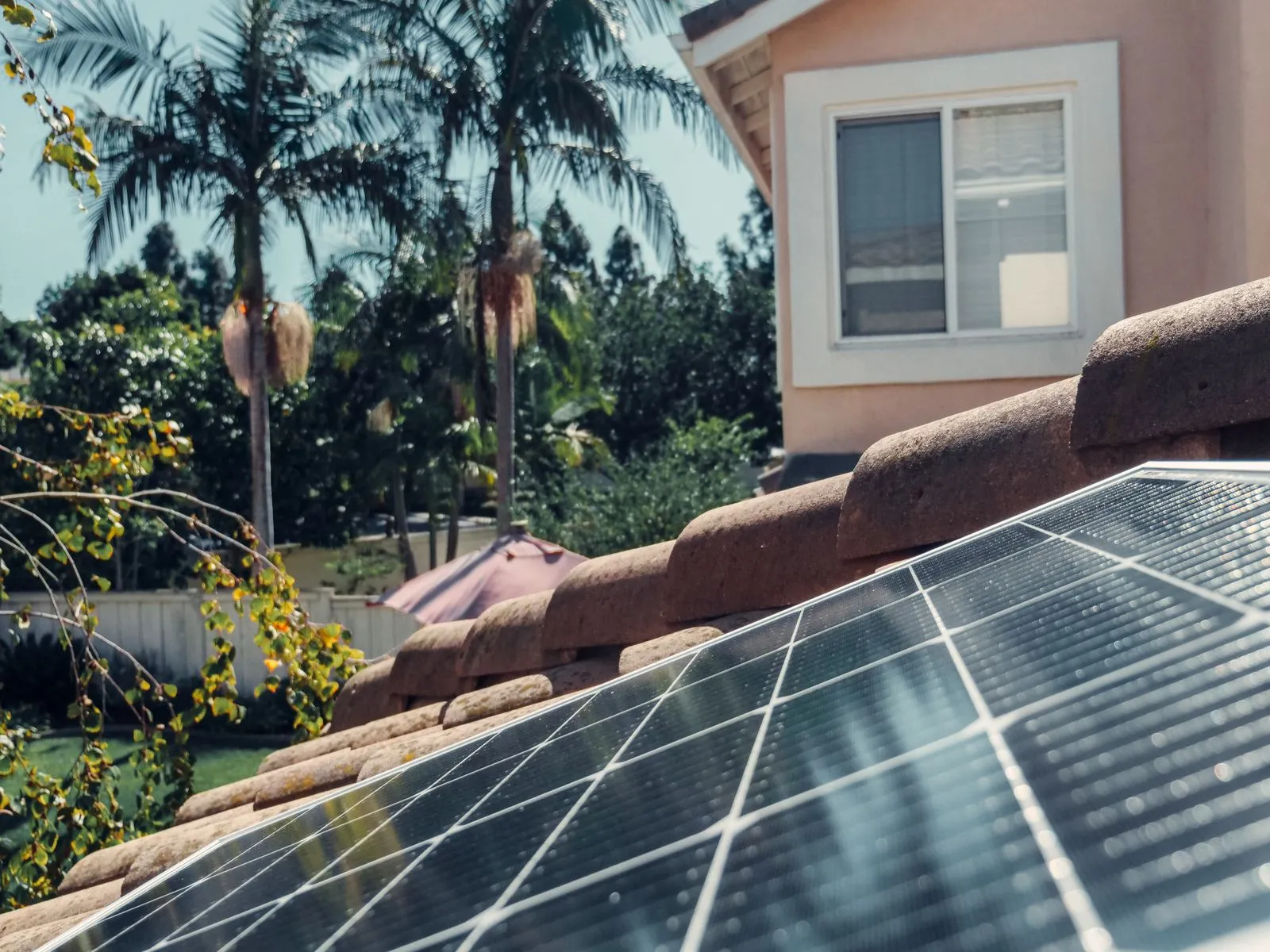 Close-up of a solar panel on a tiled roof with palm trees and a stucco house behind it