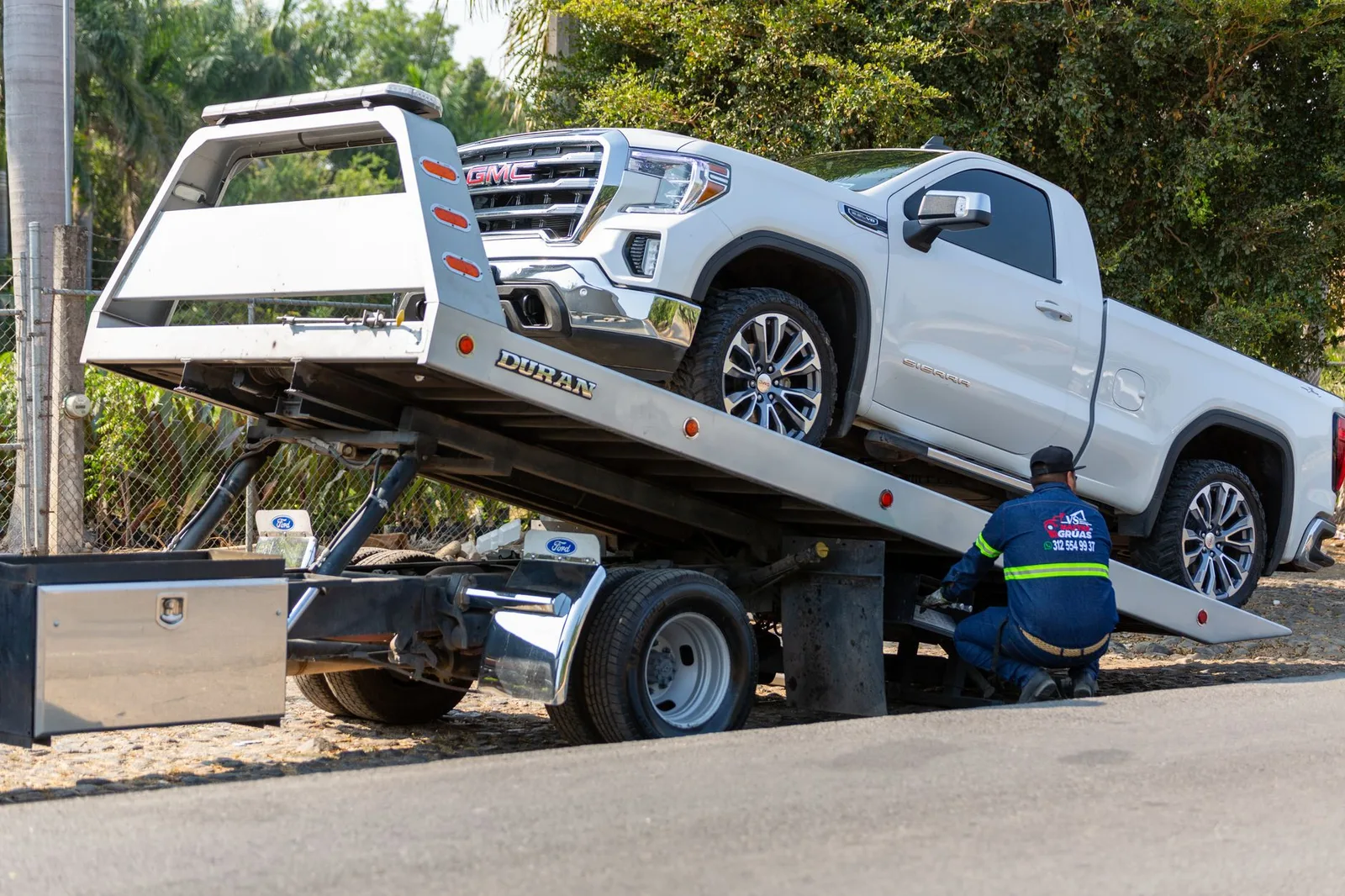 Tow truck operator loading a white pickup truck onto a flatbed on a city street