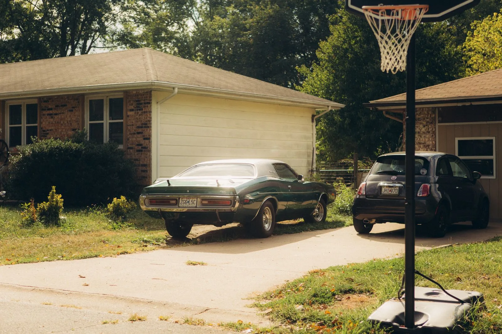 A classic car and a small hatchback parked in a suburban driveway beside a basketball hoop