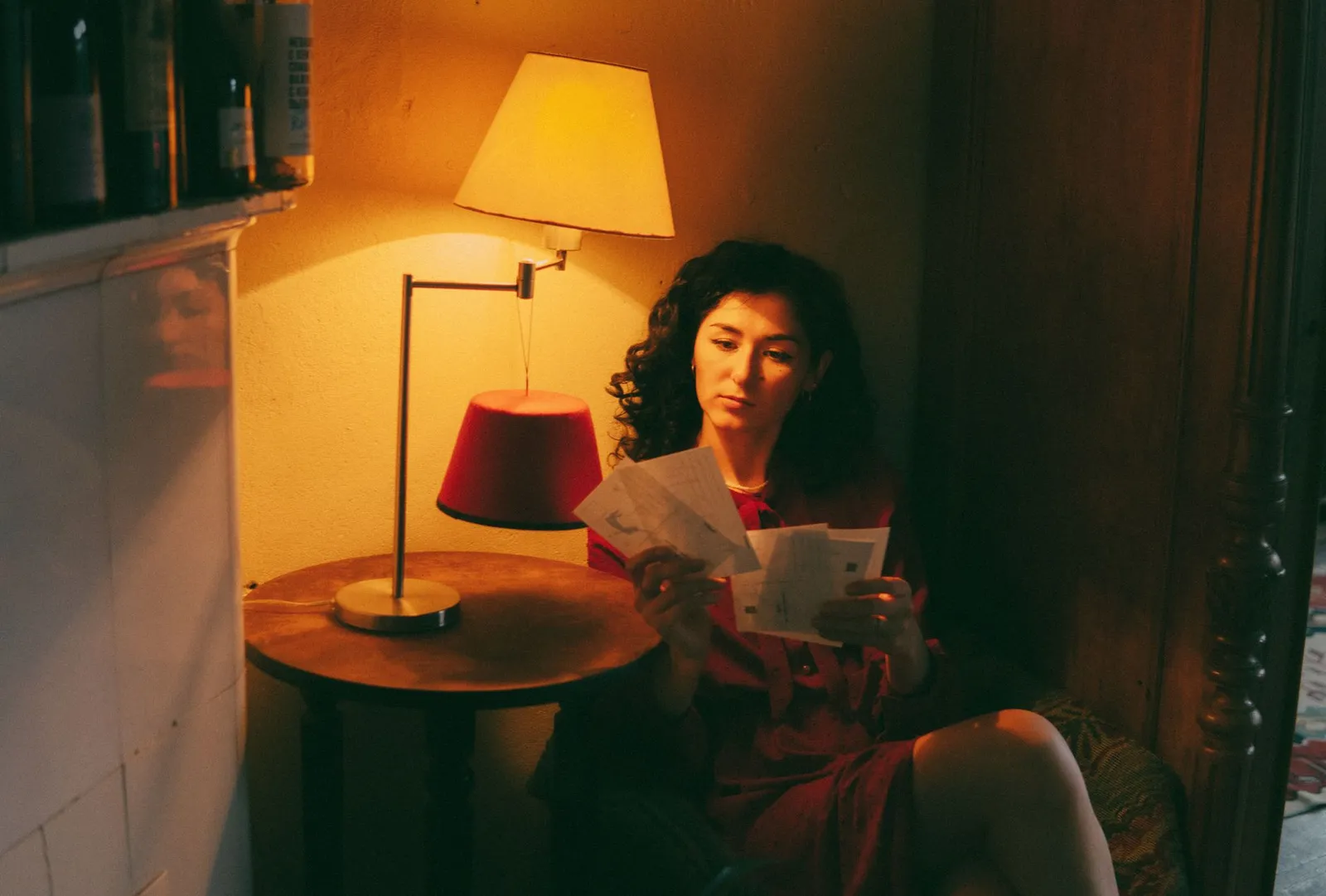 A woman sitting beside a lamp in a warmly lit room reading through a stack of letters