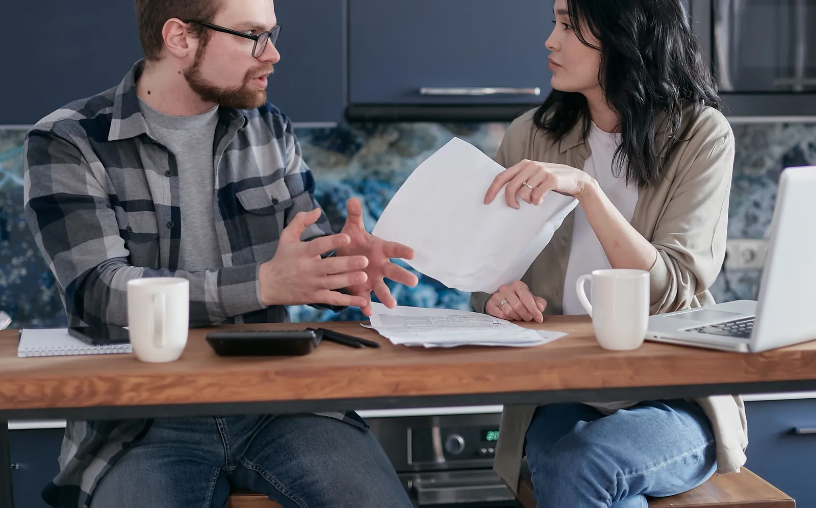 A couple at a kitchen table going through printed paperwork with a laptop, calculator, and coffee mugs