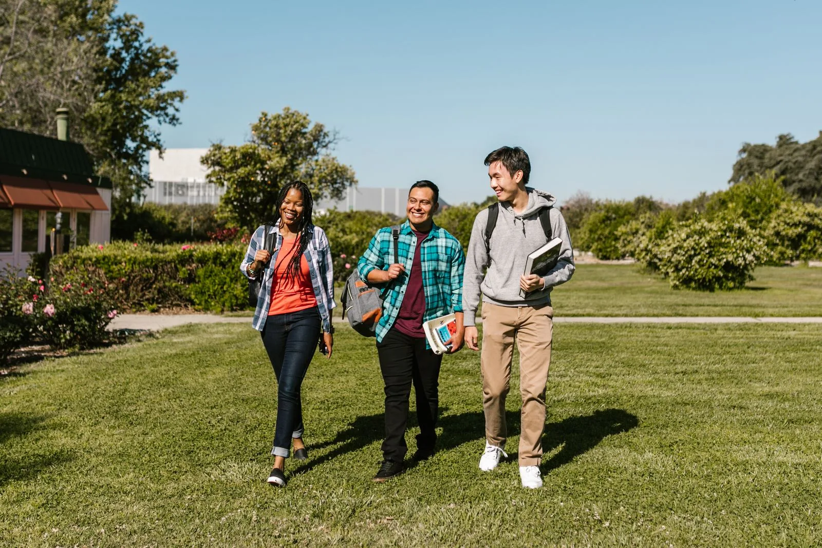 Three college students walking together across a sunlit campus lawn carrying backpacks and books