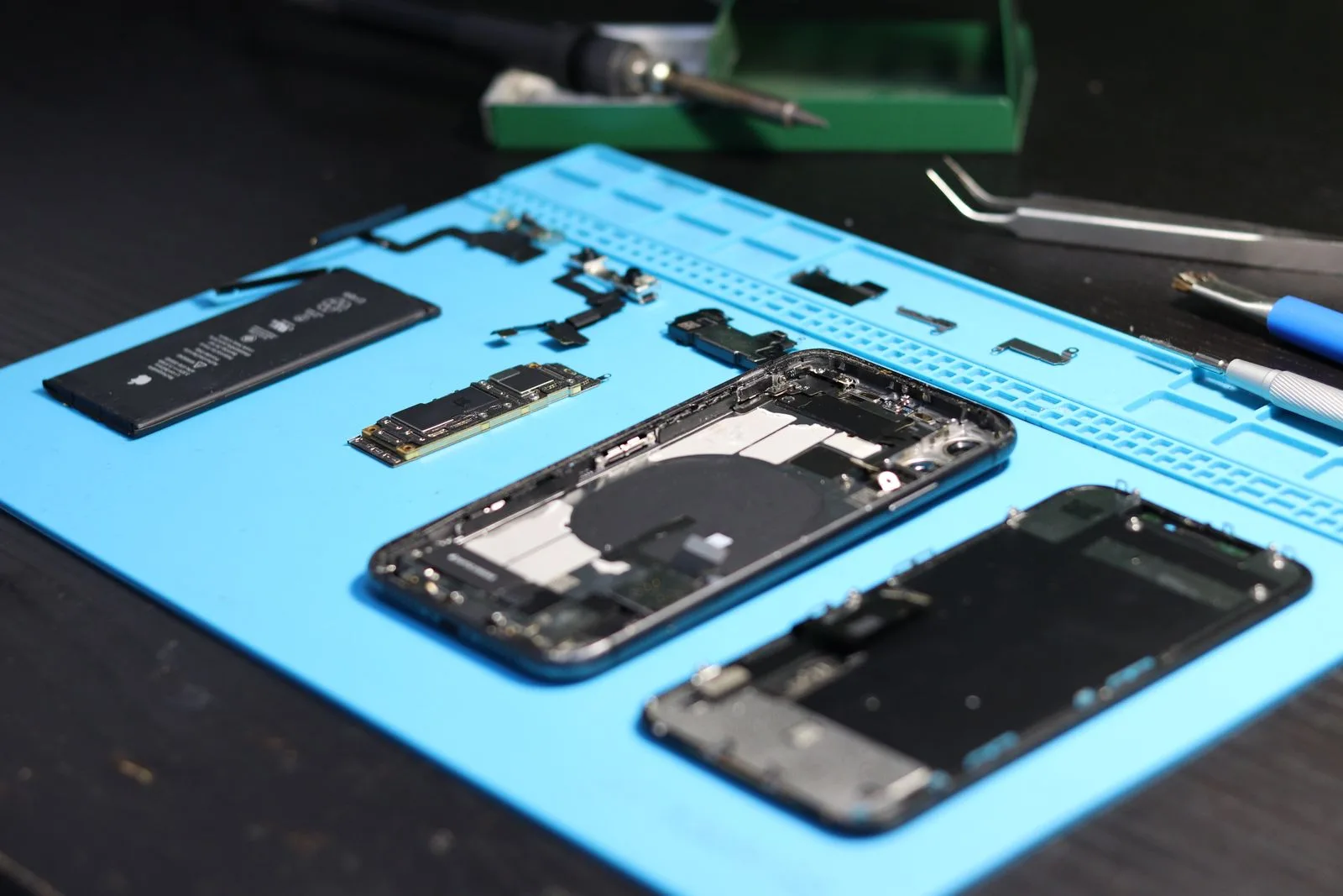 A disassembled smartphone laid out in parts on a blue repair mat beside precision tools