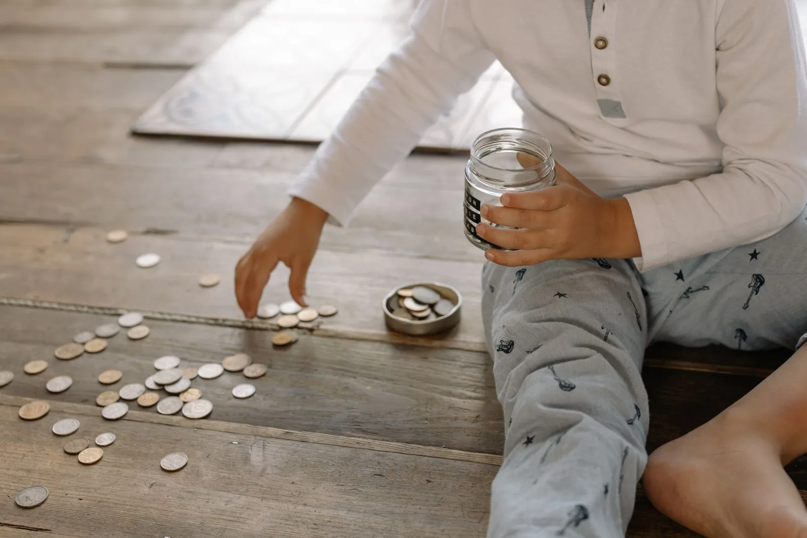 A child sitting on a wooden floor sorting coins into a glass jar