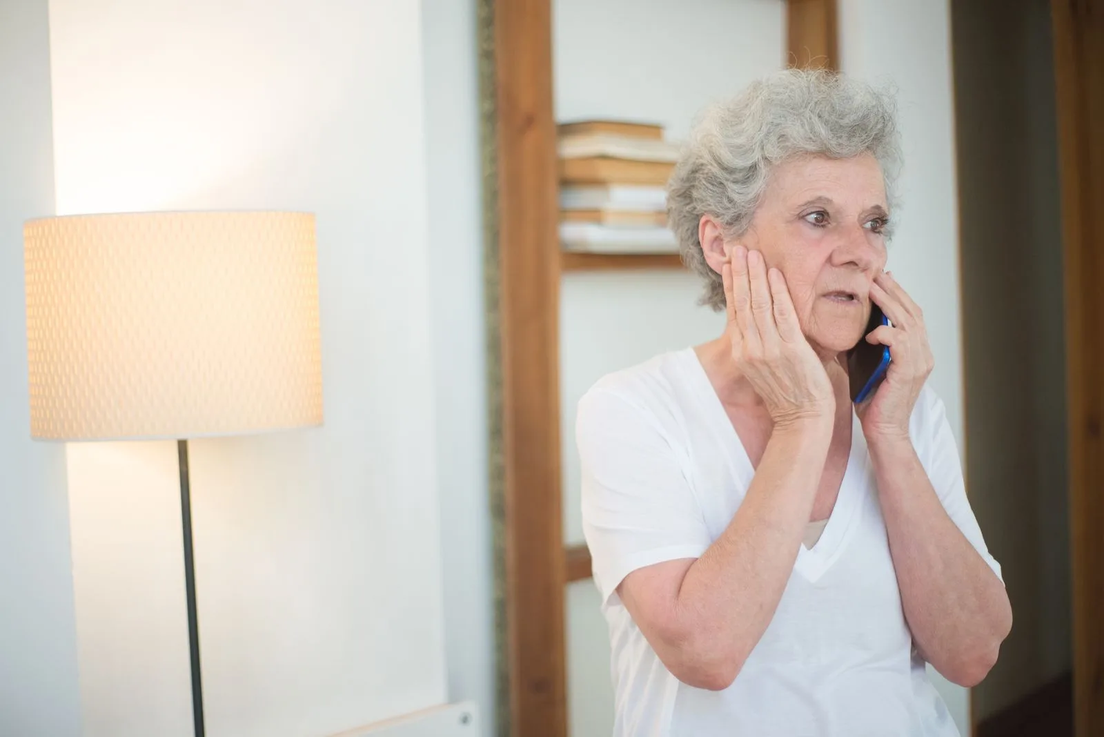 An older woman standing in her living room taking a phone call, looking worried