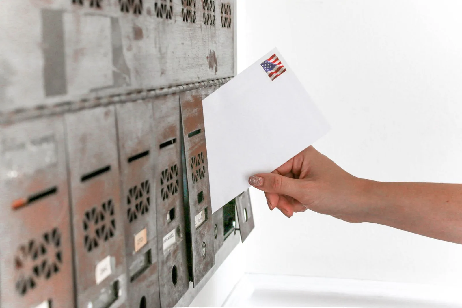 A hand posting a stamped white envelope into a bank of metal mailboxes