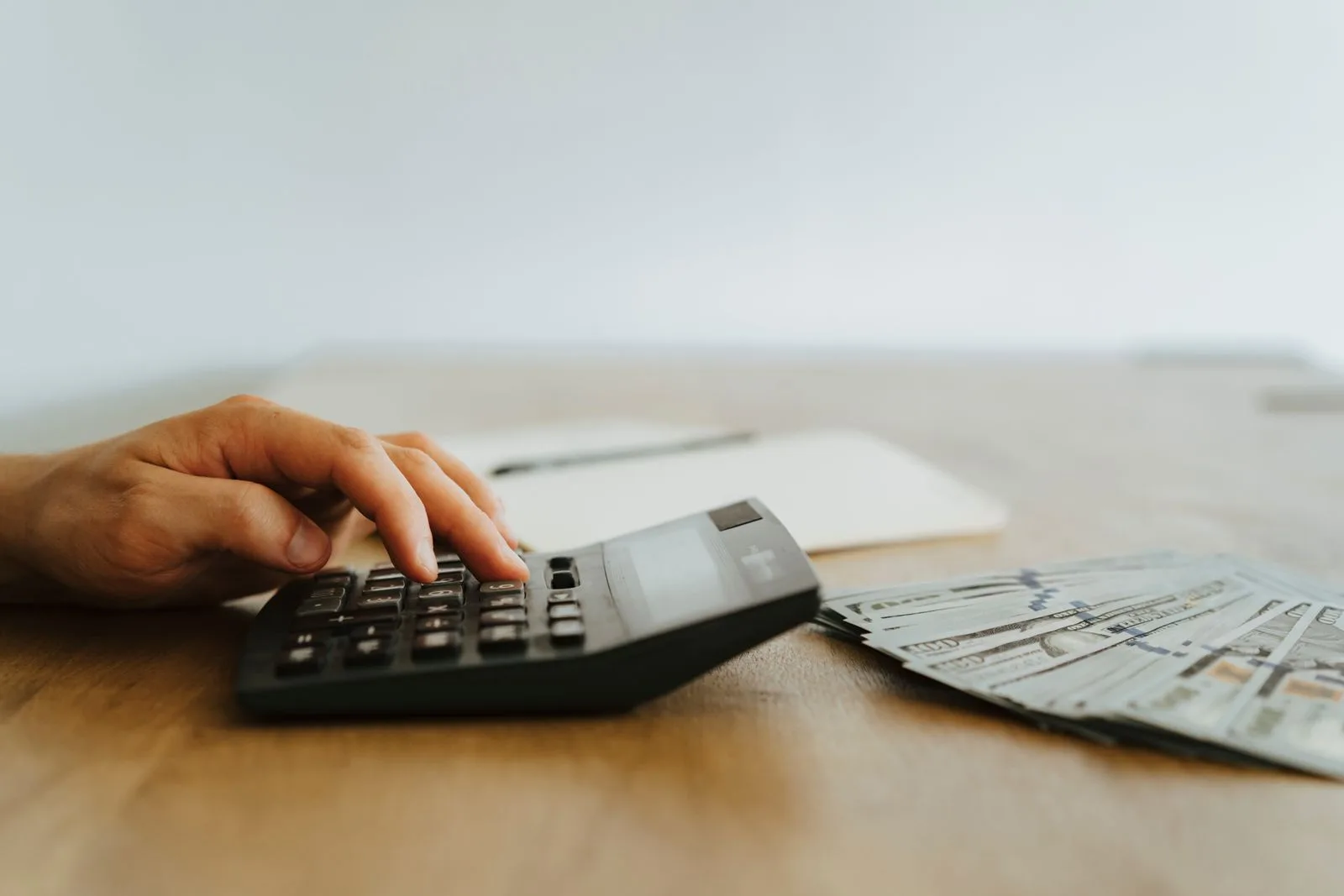 A hand using a desk calculator beside a fan of hundred dollar bills